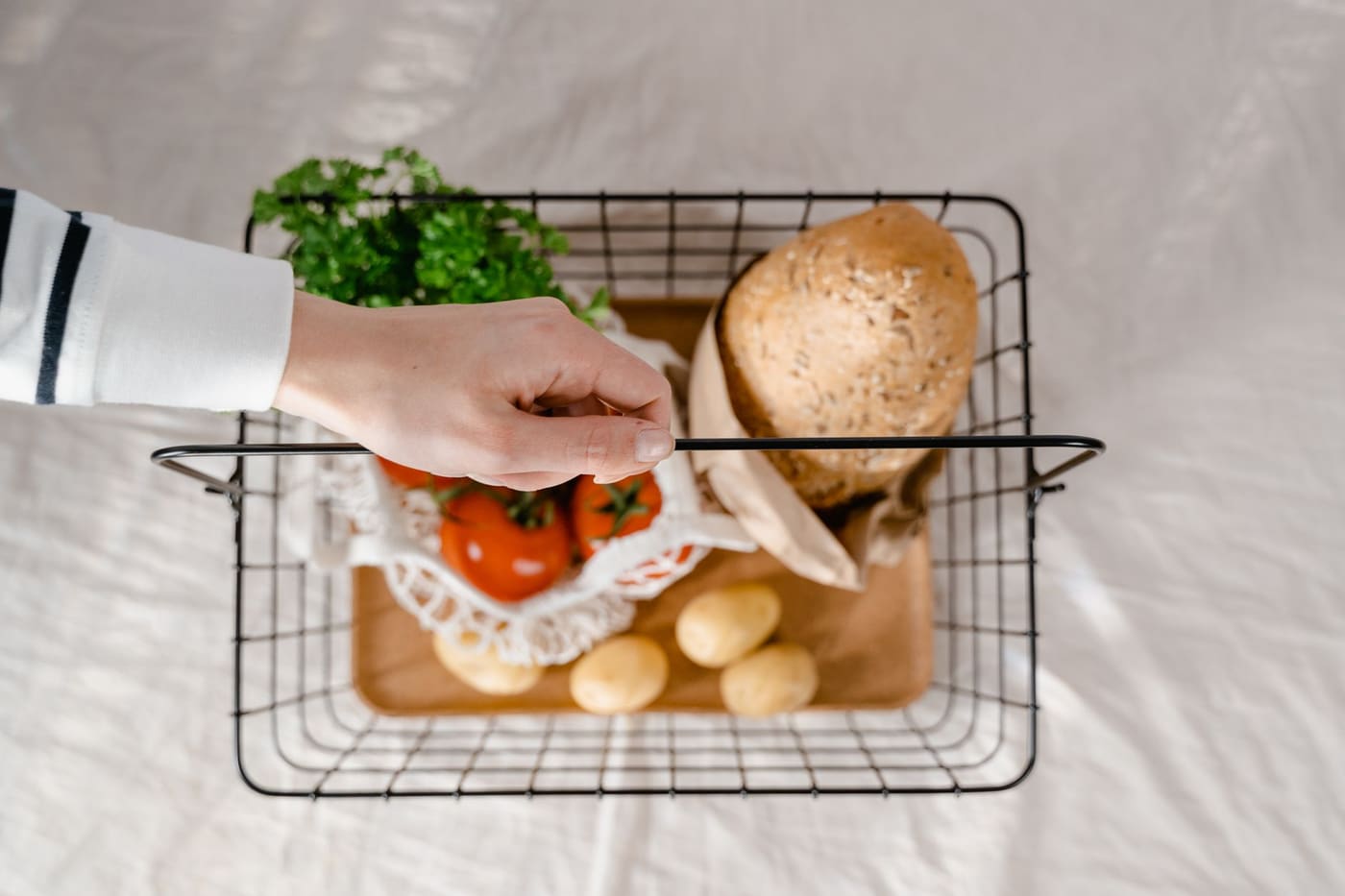 A hand holding a wire basket with bread, potatoes, tomatoes and greens