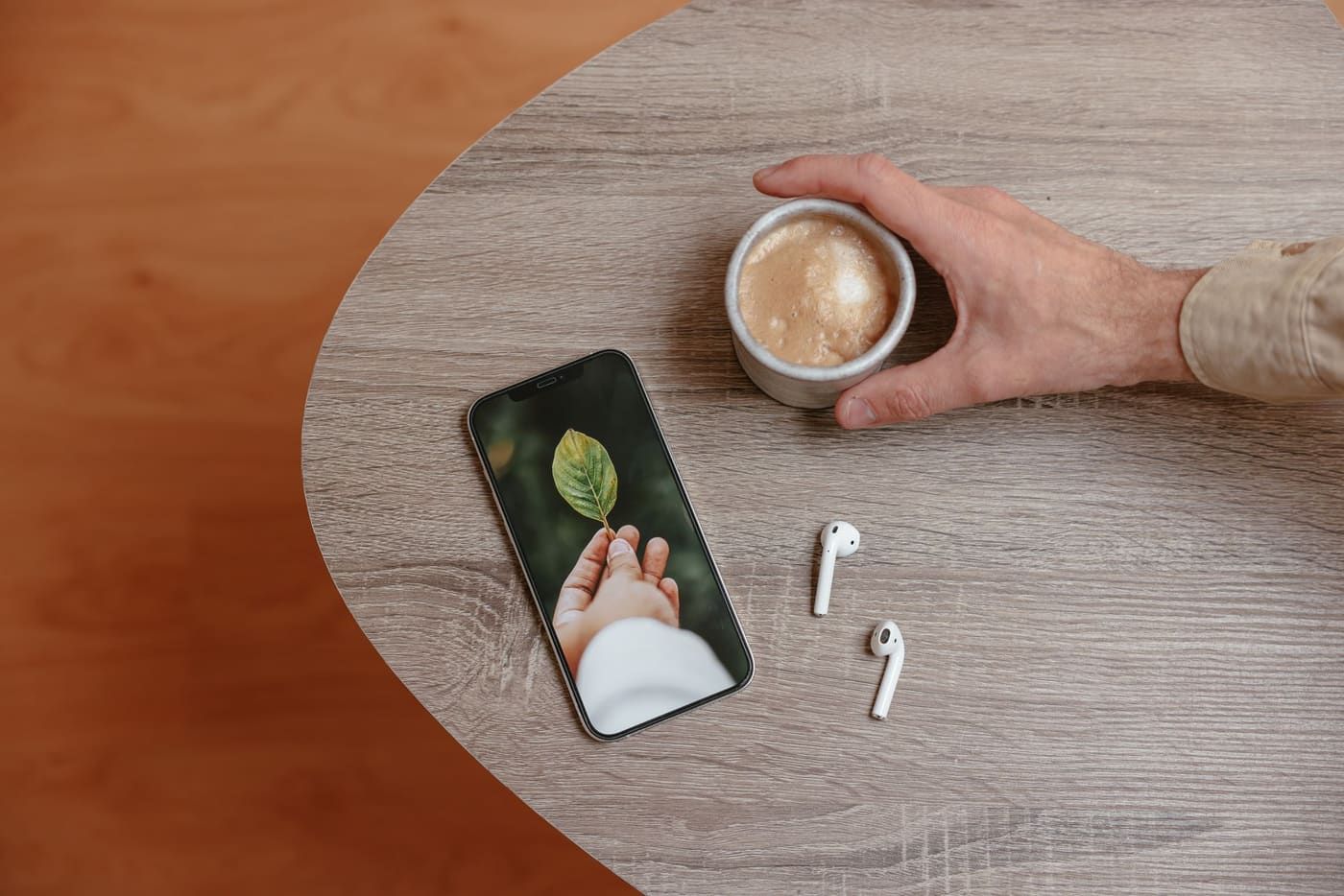 A hand holding a cup of coffee beside a smartphone and earbuds on a wooden table