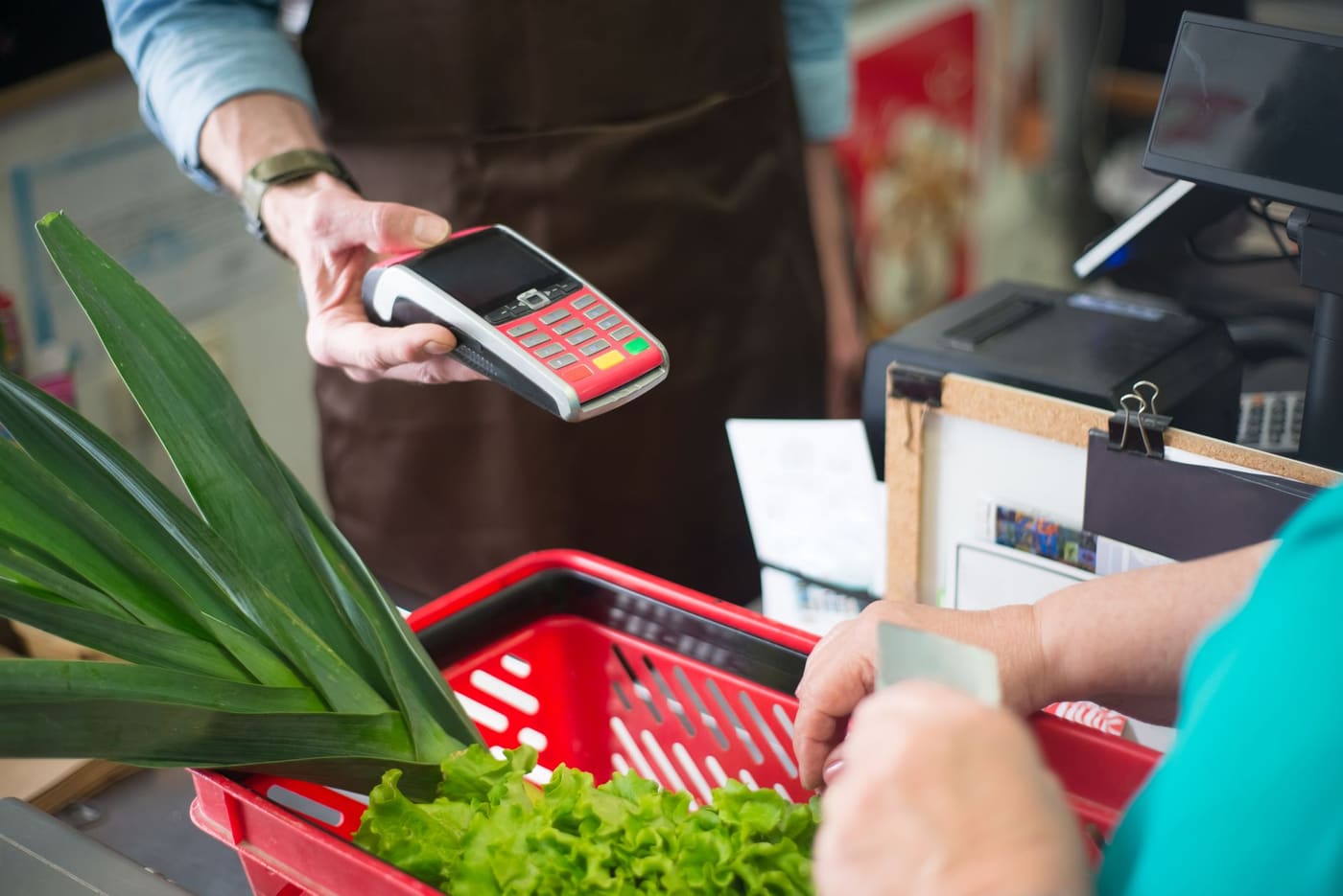 A customer paying by card at a checkout with a red basket of fresh vegetables