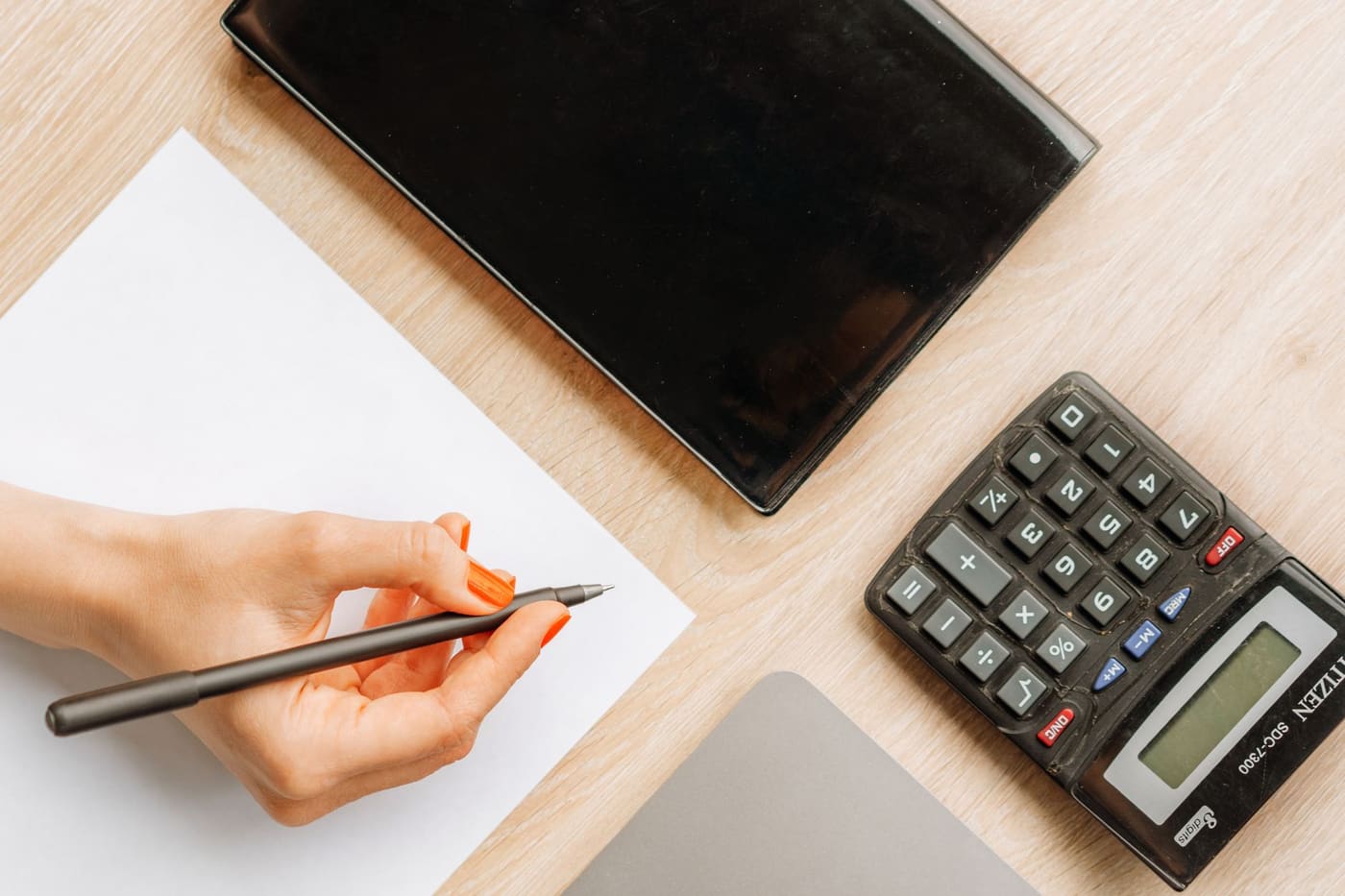 Top view of a hand writing on paper beside a calculator and a closed tablet on a wooden desk