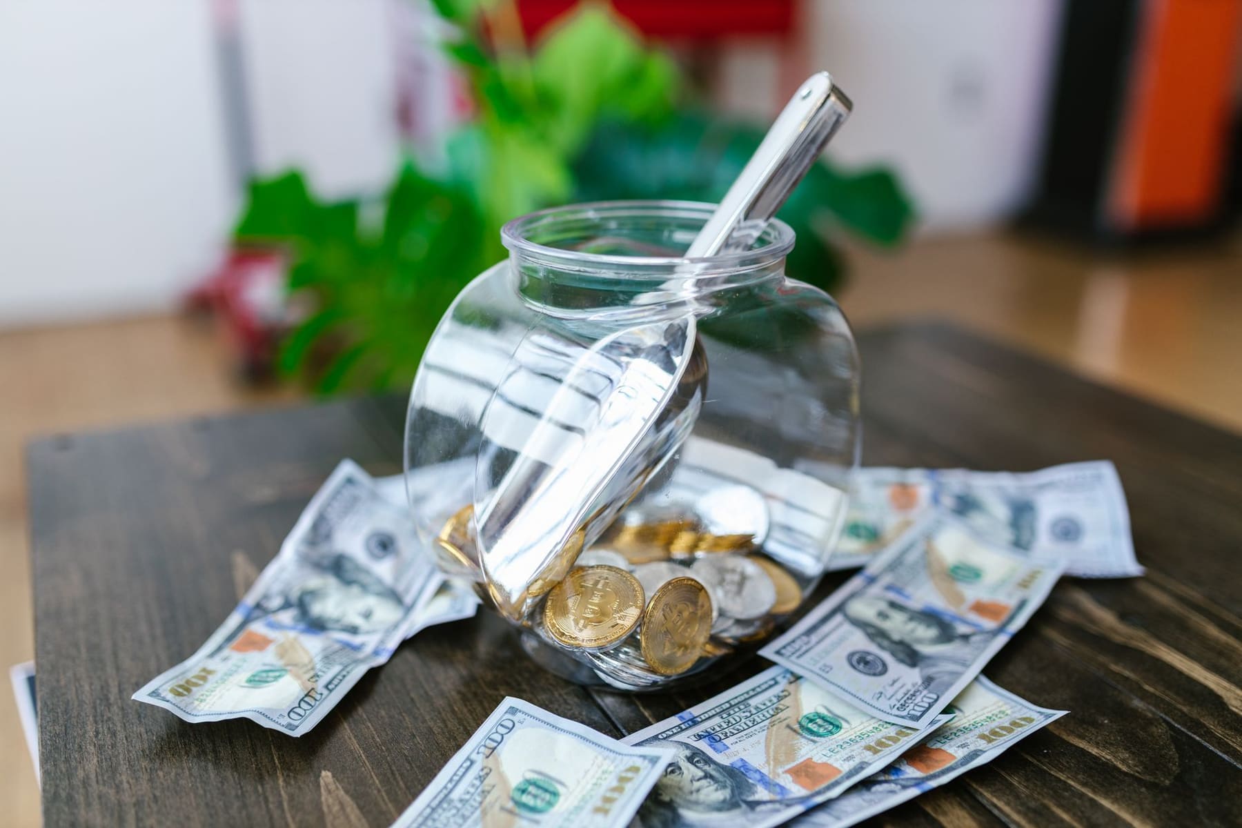A glass jar of coins with dollar bills scattered around it on a dark wooden table