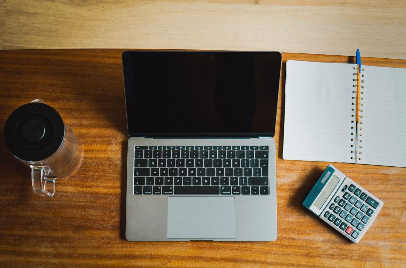 A laptop, notebook, pen and small calculator on a wooden desk seen from above