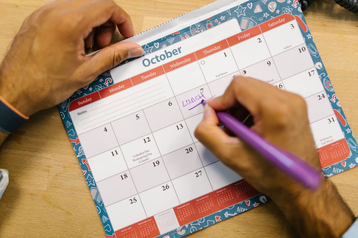 A person writing a note with a purple pen on an October wall calendar