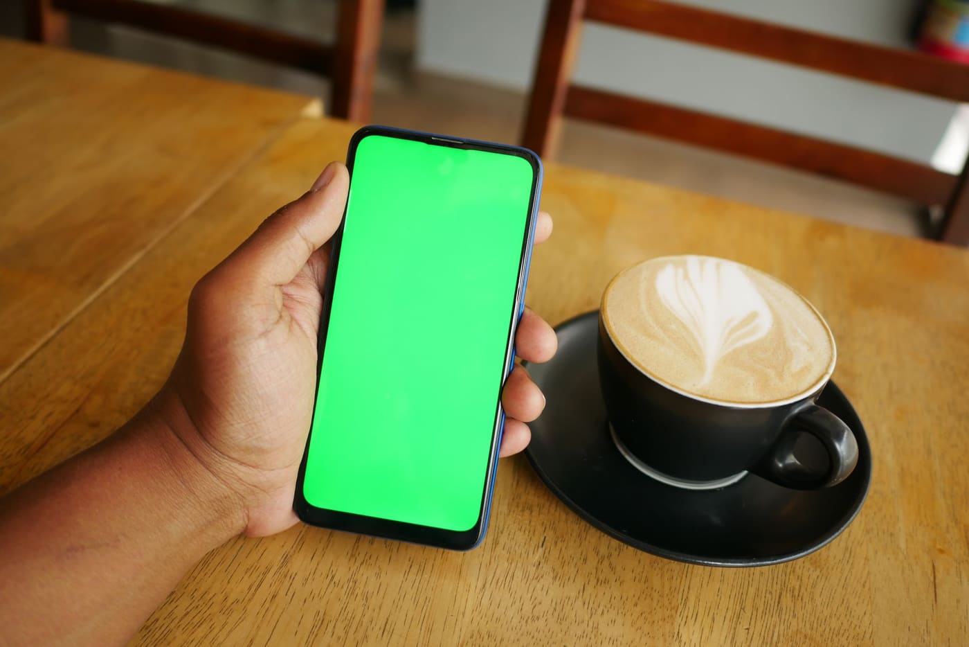 A hand holding a smartphone with a plain green screen next to a latte on a wooden table