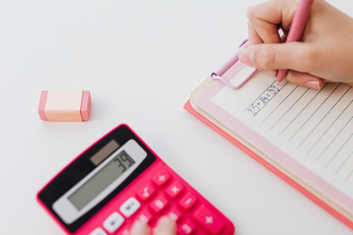 A hand writing in a notebook next to a pink calculator on a white desk